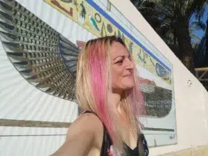 Mujer de perfil, sonriente, apoyada en un muro que tiene pintado un mural con la diosa egipcia Isis, con sus grandes alas desplegadas y una cenefa con símbolos típicos egipcios, antiguos