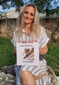 Mujer sentada, en un entorno natural, con césped y un muro de piedra antigua en el fondo, que sostiene un libro cuadrado, titulado: De la sensibilidad a las palabras. Se muestra sonriente.